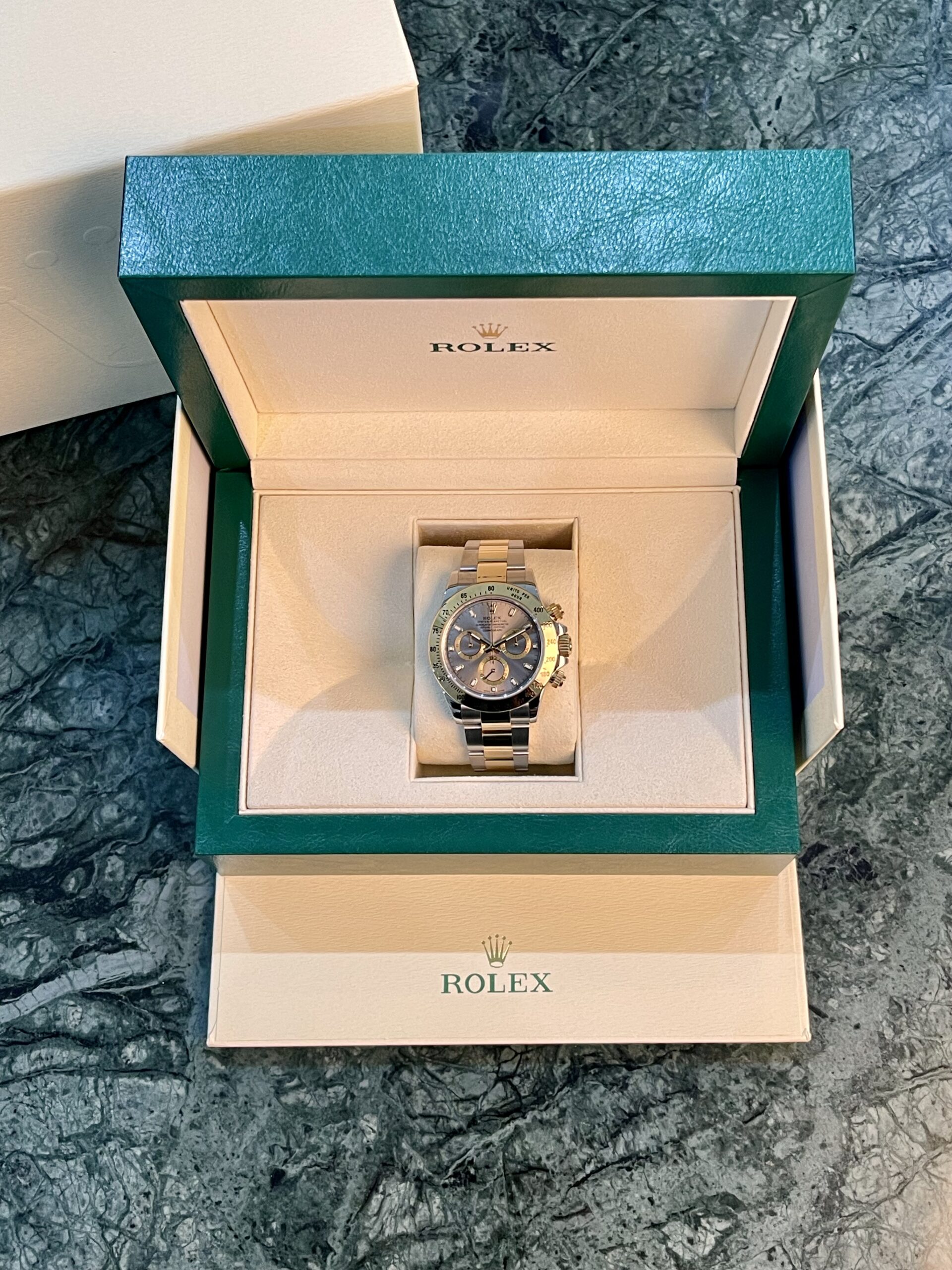 Open Rolex presentation box with a two-tone Rolex watch on a cushioned insert; green outer box and logo visible on marble surface