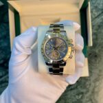 Close-up of a Rolex stainless steel chronograph watch on a white display cushion, held by gloved hands inside a green presentation box.