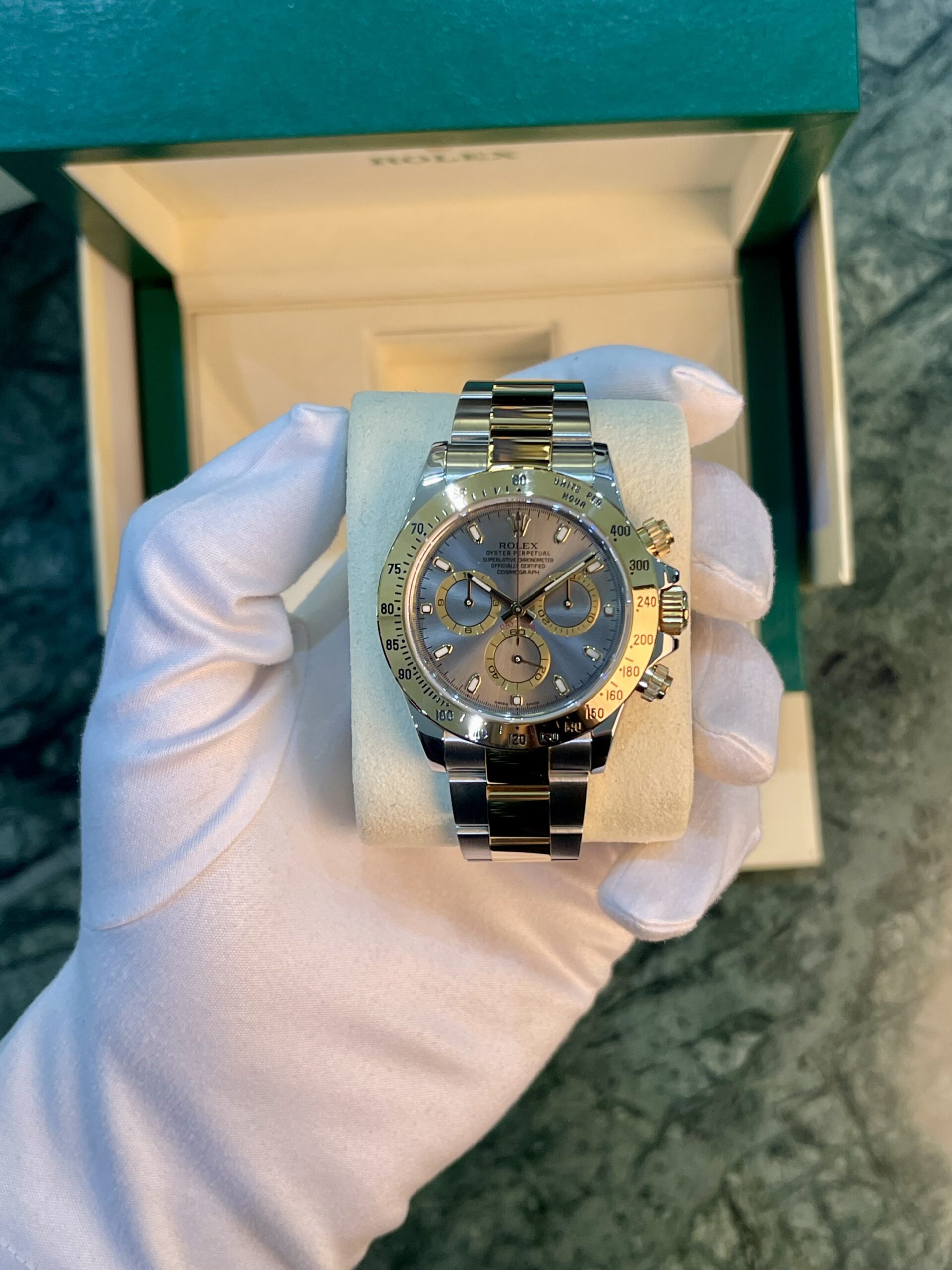 Close-up of a Rolex stainless steel chronograph watch on a white display cushion, held by gloved hands inside a green presentation box.