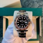 Rolex GMT‑Master II stainless steel watch with black dial and brown/beige 24‑hour bezel on a light cushion inside a green presentation box.