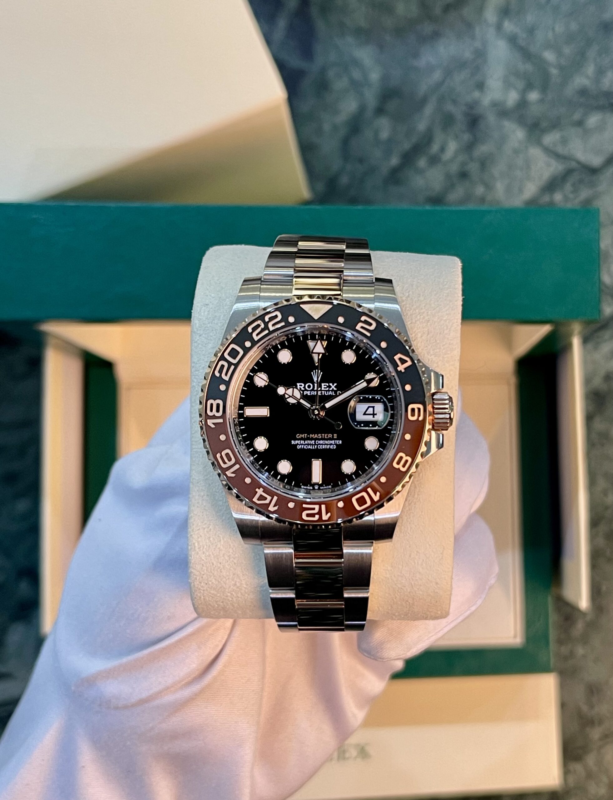 Rolex GMT‑Master II stainless steel watch with black dial and brown/beige 24‑hour bezel on a light cushion inside a green presentation box.