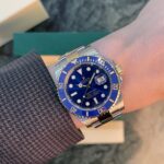 Close-up of a two-tone Rolex Submariner with a blue dial and blue bezel on a wrist, worn over a textured blazer sleeve.