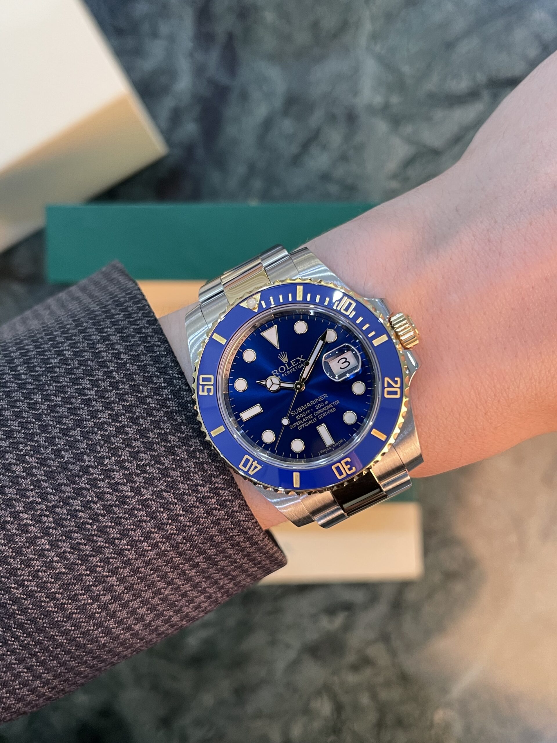 Close-up of a two-tone Rolex Submariner with a blue dial and blue bezel on a wrist, worn over a textured blazer sleeve.
