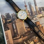 Gold wristwatch with white dial and black leather strap resting on a Shanghai skyline poster/photograph.