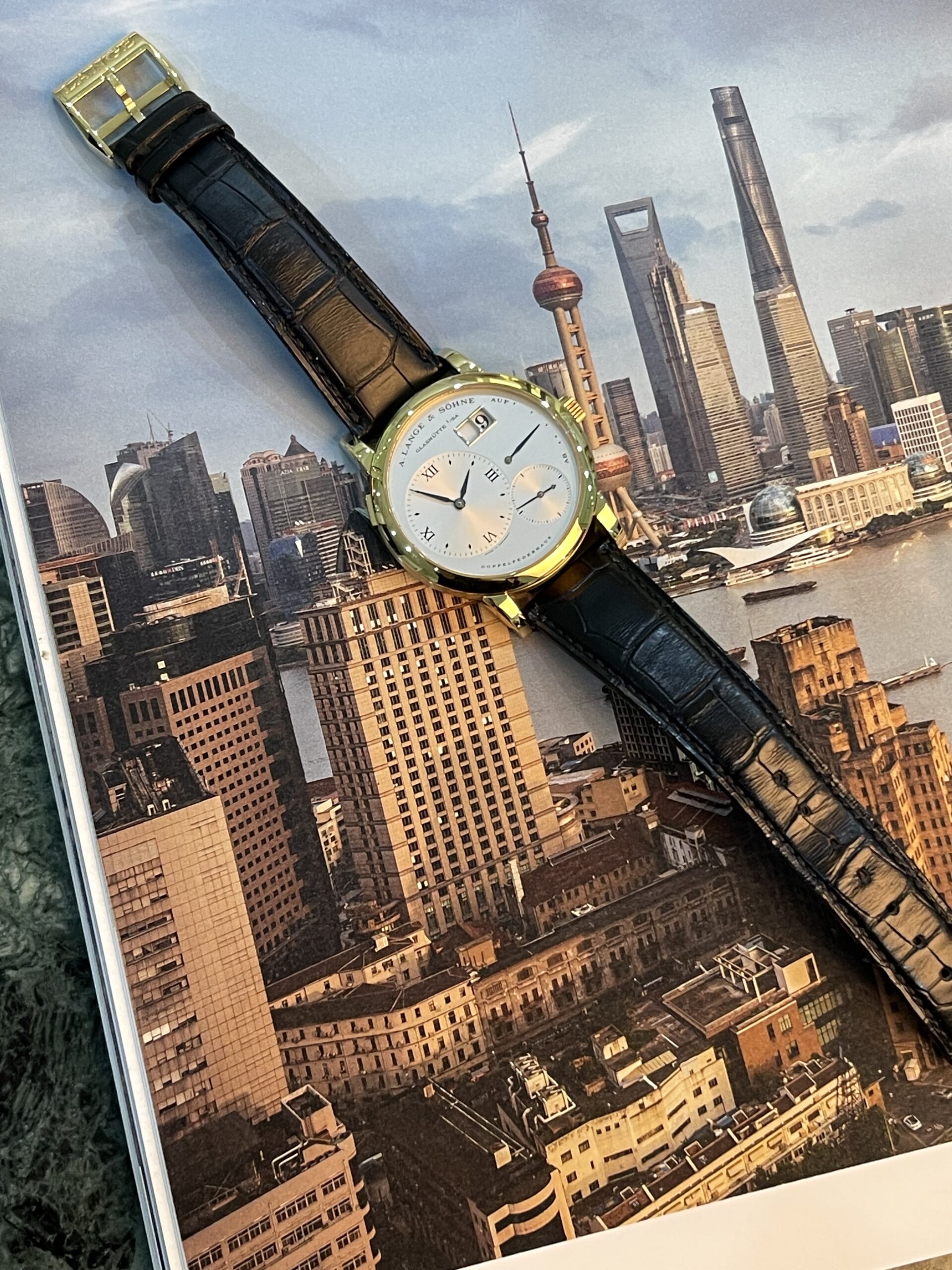 Gold wristwatch with white dial and black leather strap resting on a Shanghai skyline poster/photograph.
