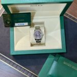 Green Rolex box open to reveal a silver watch on a cream cushion, with a warranty card and booklets nearby.