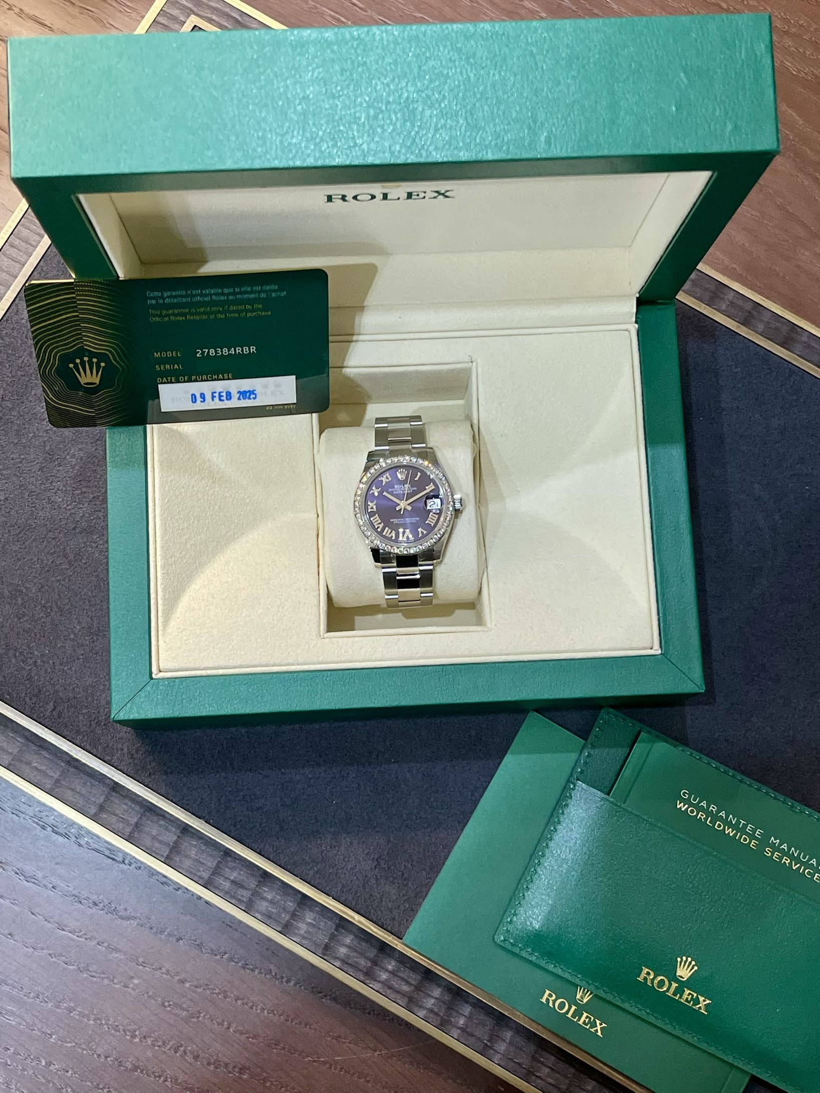 Green Rolex box open to reveal a silver watch on a cream cushion, with a warranty card and booklets nearby.