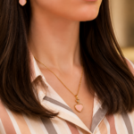 Close-up of a woman wearing a gold heart pendant necklace and matching heart earring, with a striped blouse visible in the background.