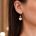 Close-up of a woman's ear wearing a gold hoop earring with two heart charms: a pink lower heart and a small gray upper heart.