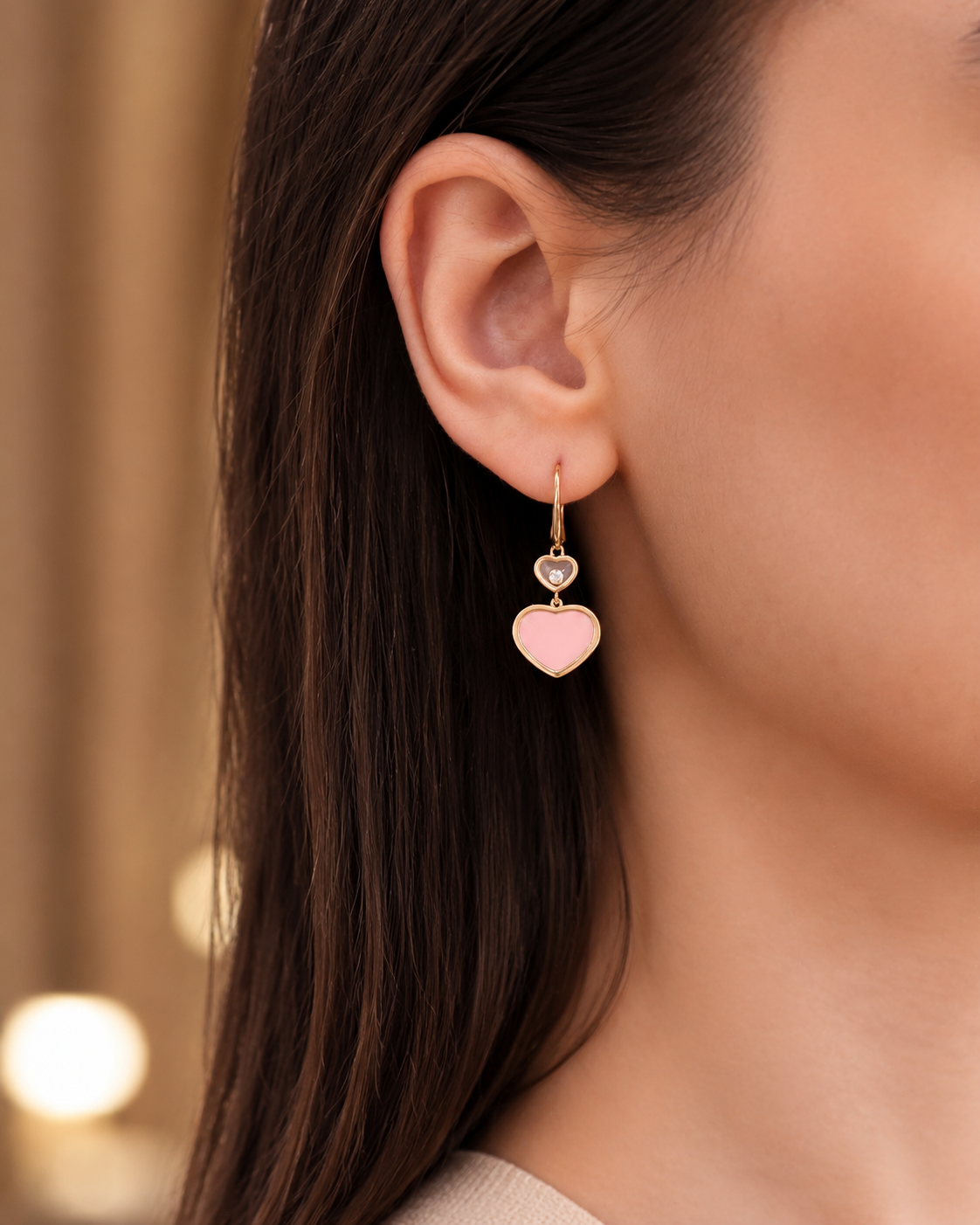 Close-up of a woman's ear wearing a gold hoop earring with two heart charms: a pink lower heart and a small gray upper heart.