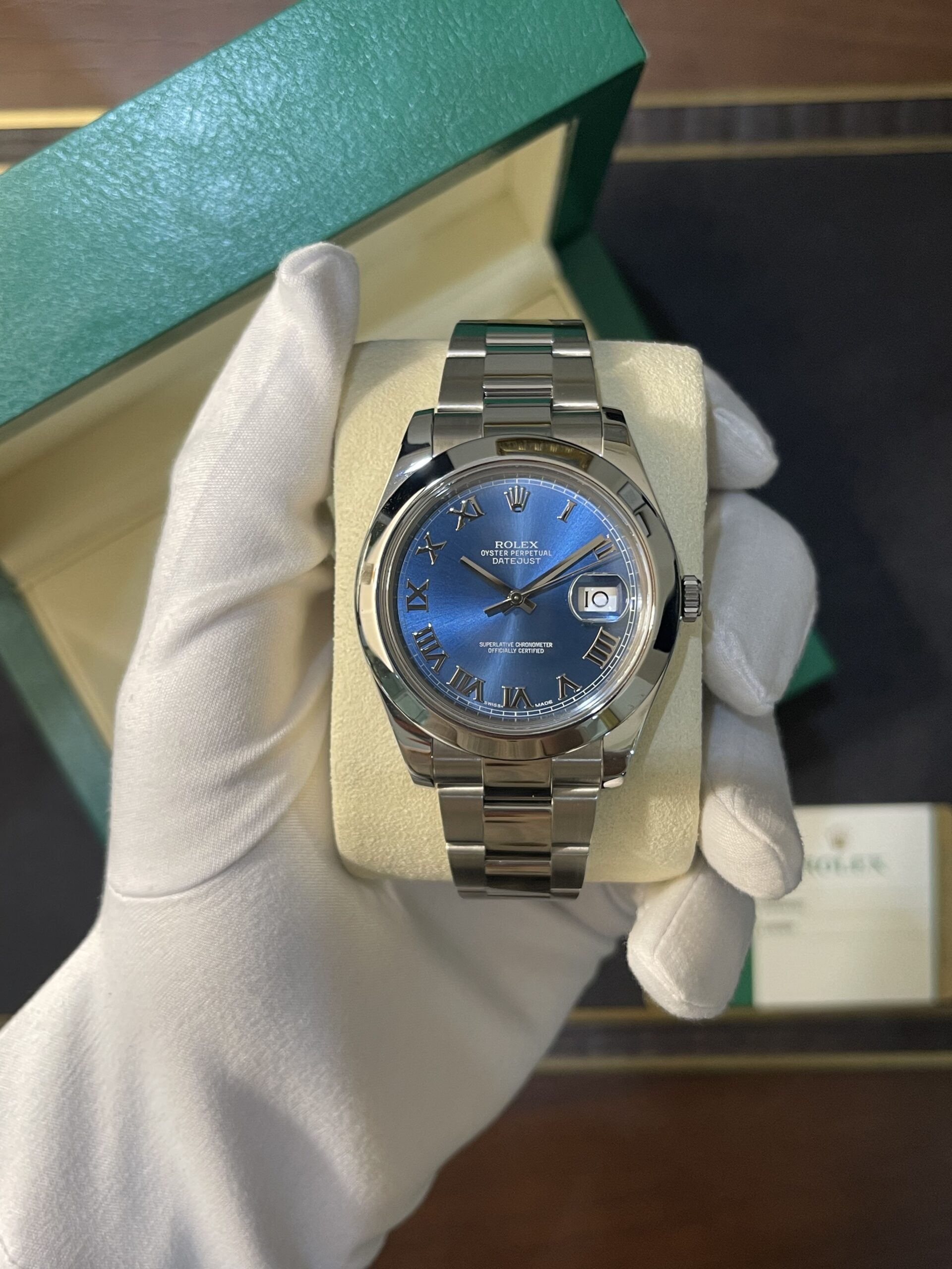Rolex Datejust watch with blue dial and Roman numeral markers in a green presentation box held by gloved hand