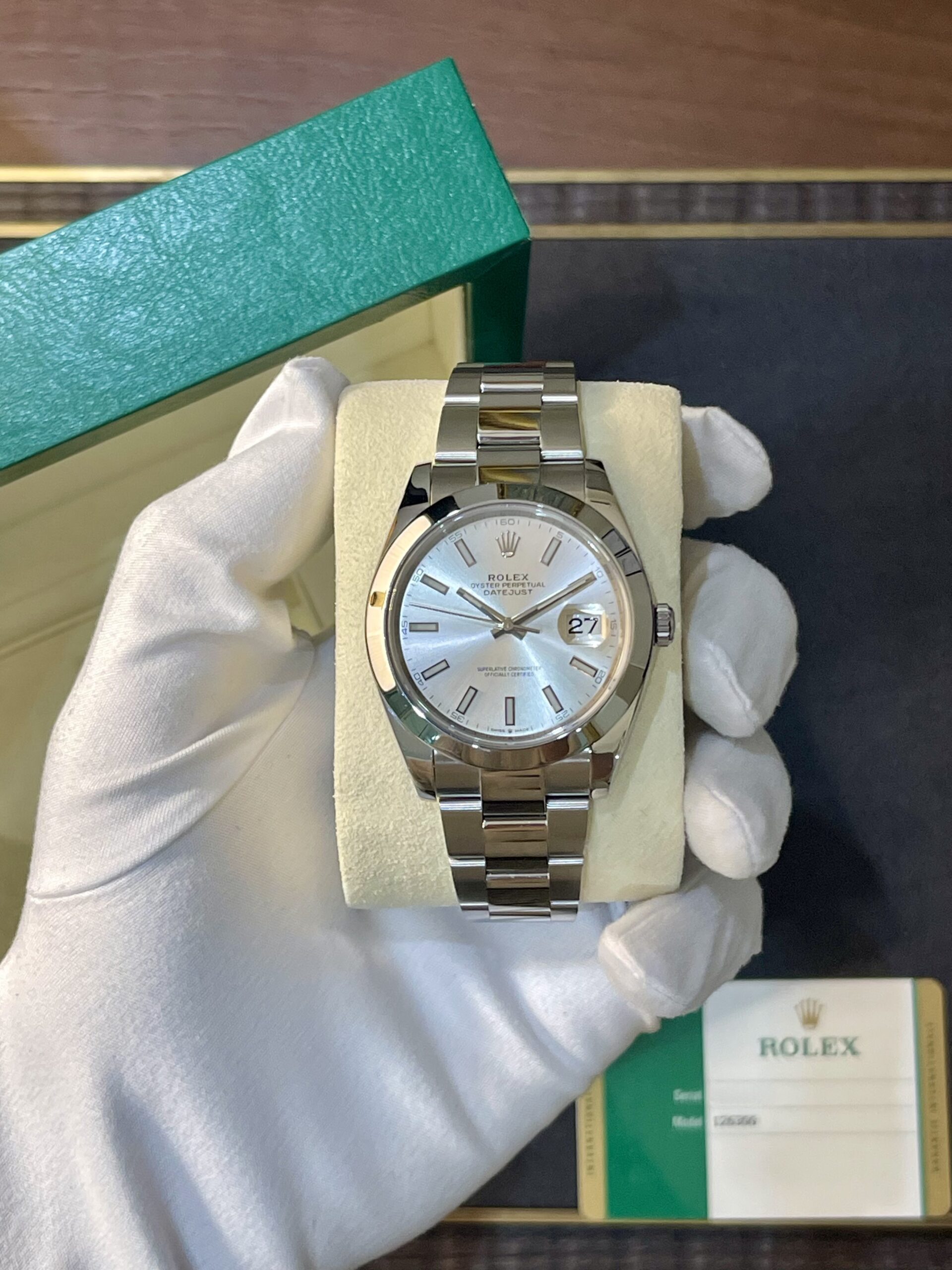Silver Rolex Datejust wristwatch with a metal bracelet displayed on a cream cushion inside an open green Rolex box, held by gloved hand