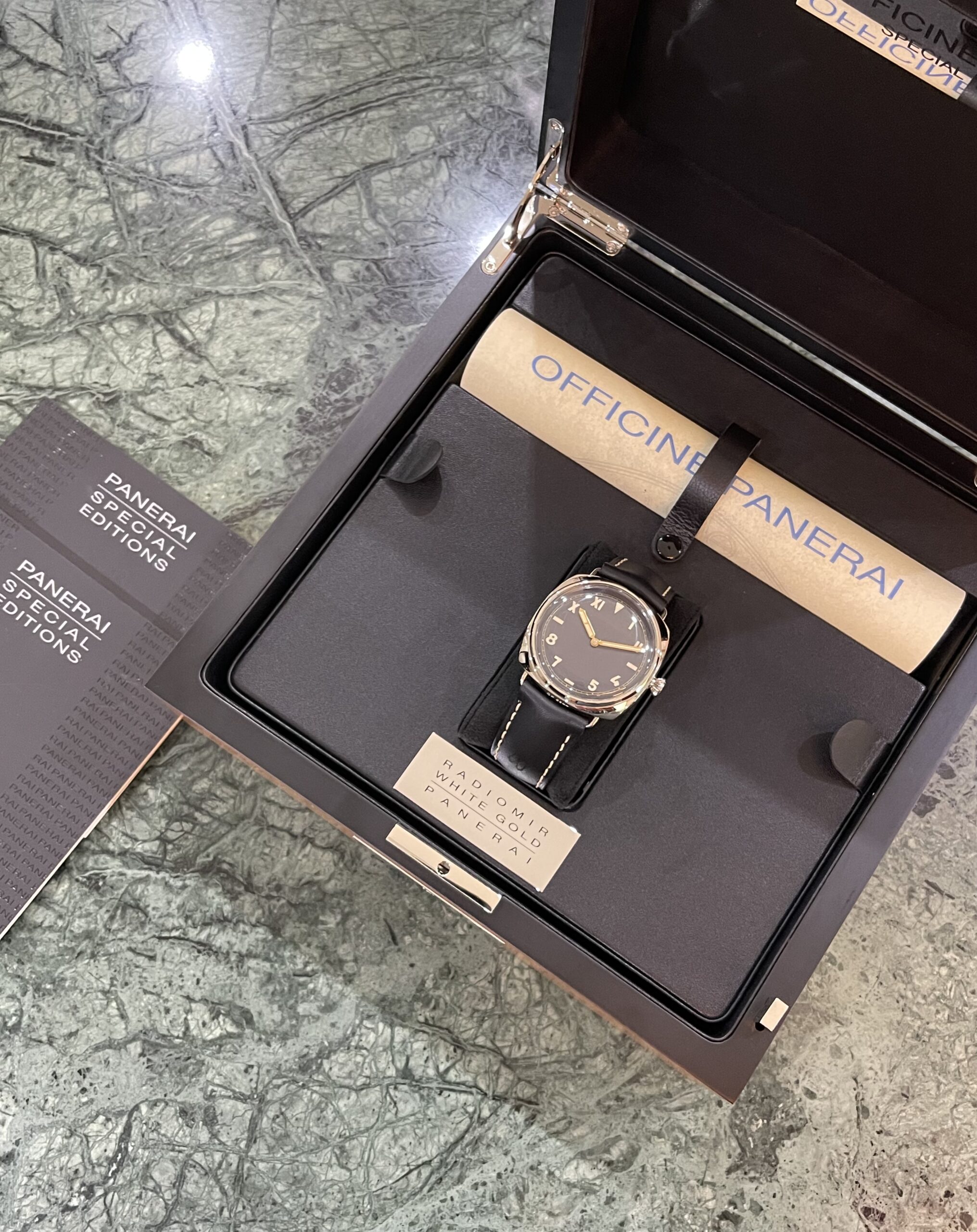 Panerai watch in an open presentation box with a black dial and gold case, brown leather strap, beside Panerai edition booklets.