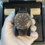 Watch with black dial and tan numerals on a black leather strap held by gloved hand inside a presentation box.