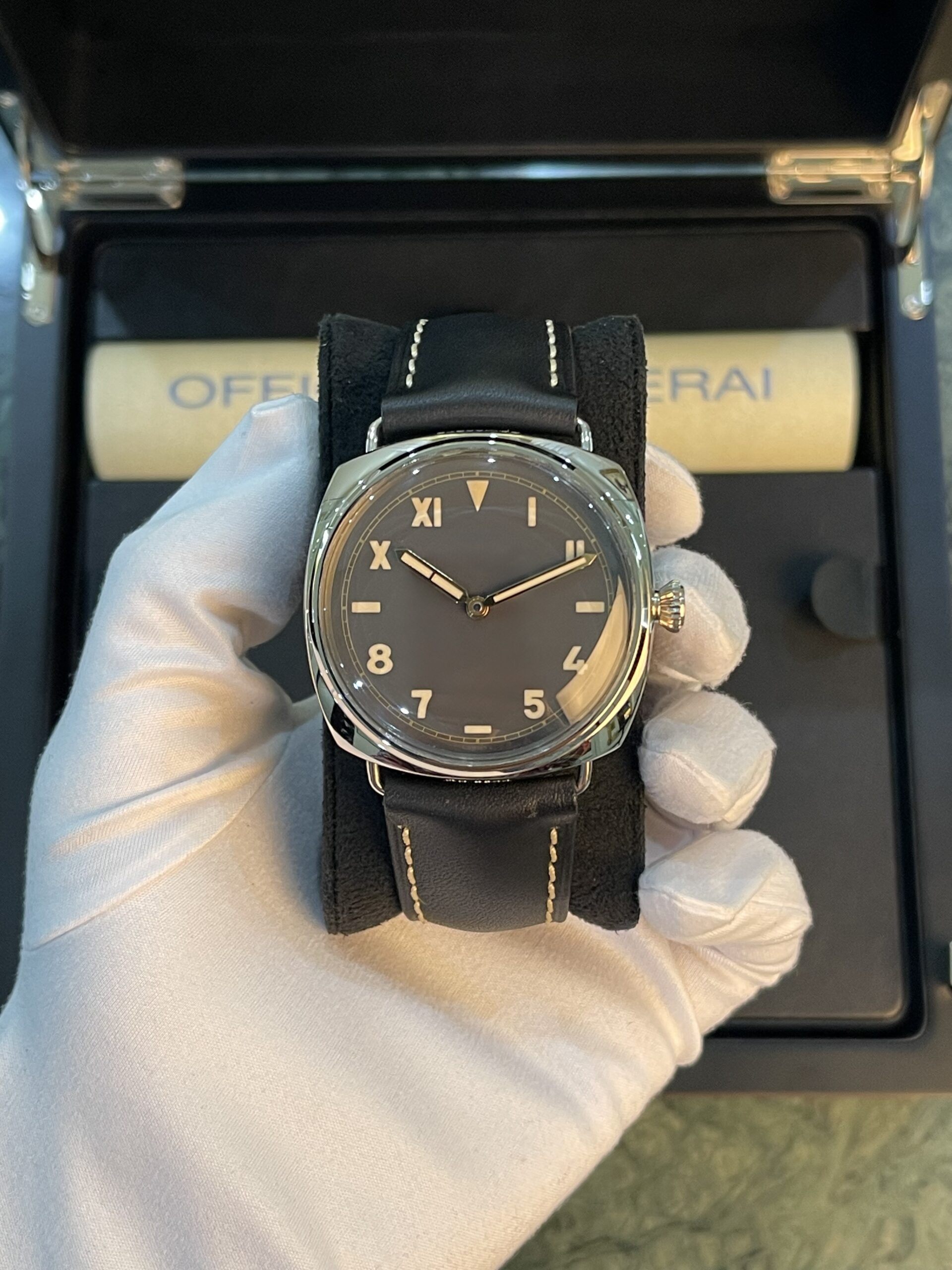 Watch with black dial and tan numerals on a black leather strap held by gloved hand inside a presentation box.