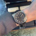 Wristwatch with a black dial and beige numerals on a leather strap, worn over a textured jacket sleeve inside a presentation box labeled Officine Panerai