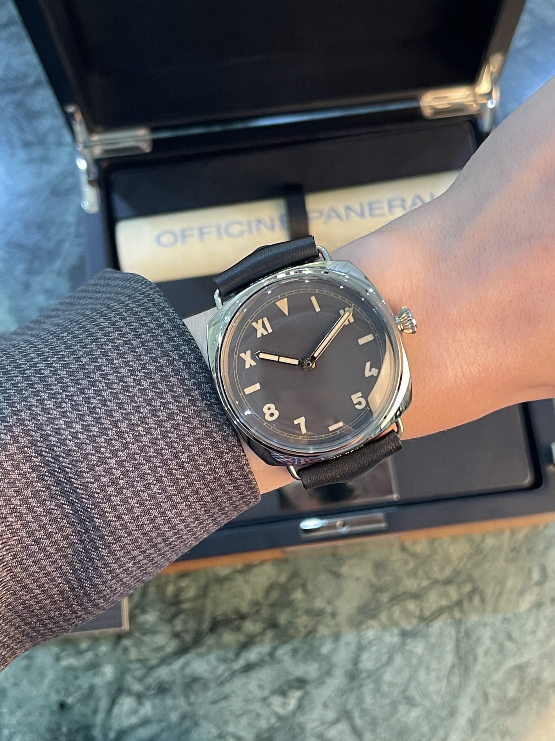Wristwatch with a black dial and beige numerals on a leather strap, worn over a textured jacket sleeve inside a presentation box labeled Officine Panerai