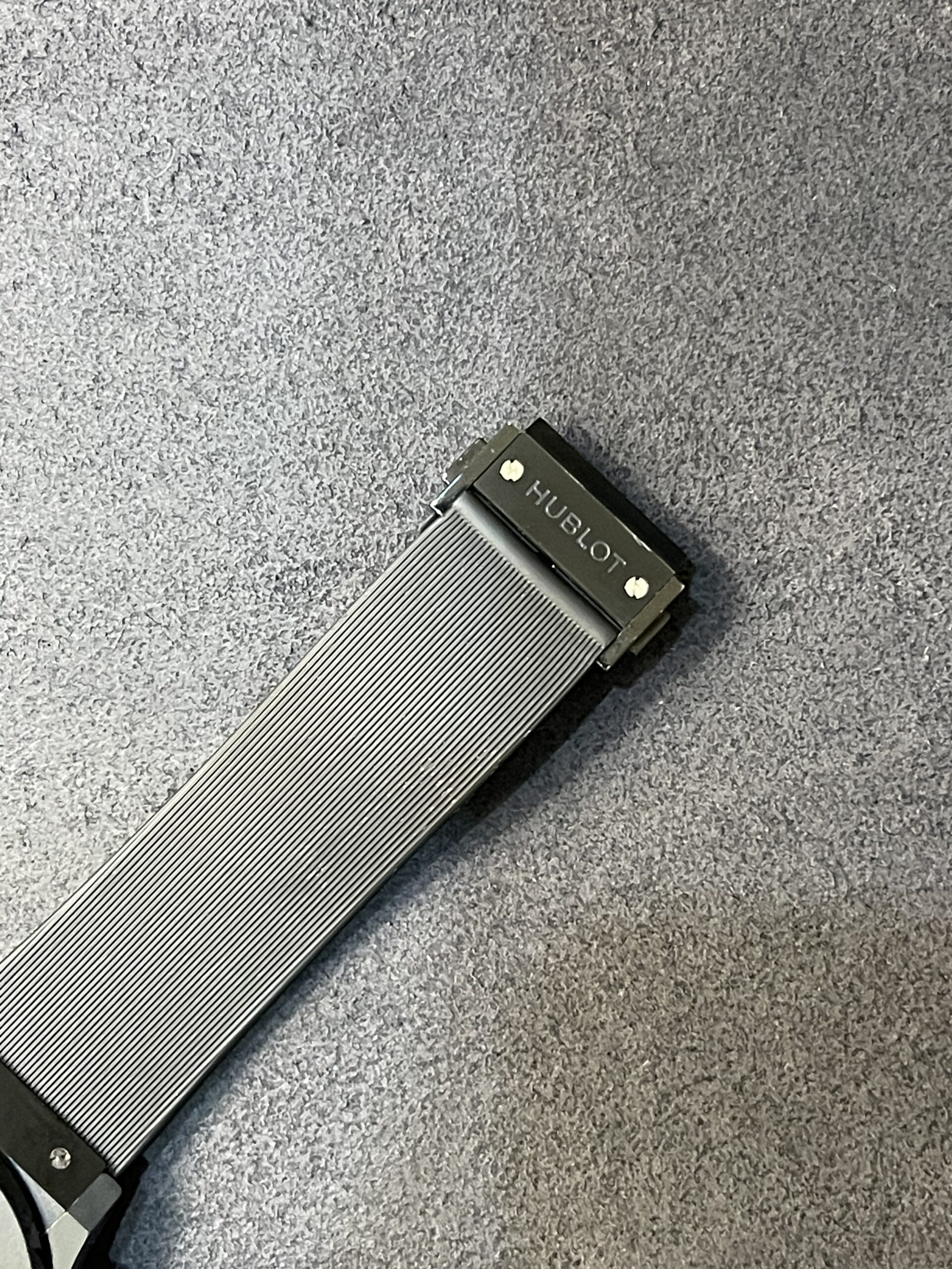Close-up of a black metal watch band with Hublot clasp against a gray carpet.