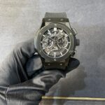 Skeleton-dial black Hublot automatic wristwatch on a gloved hand against a gray backdrop.