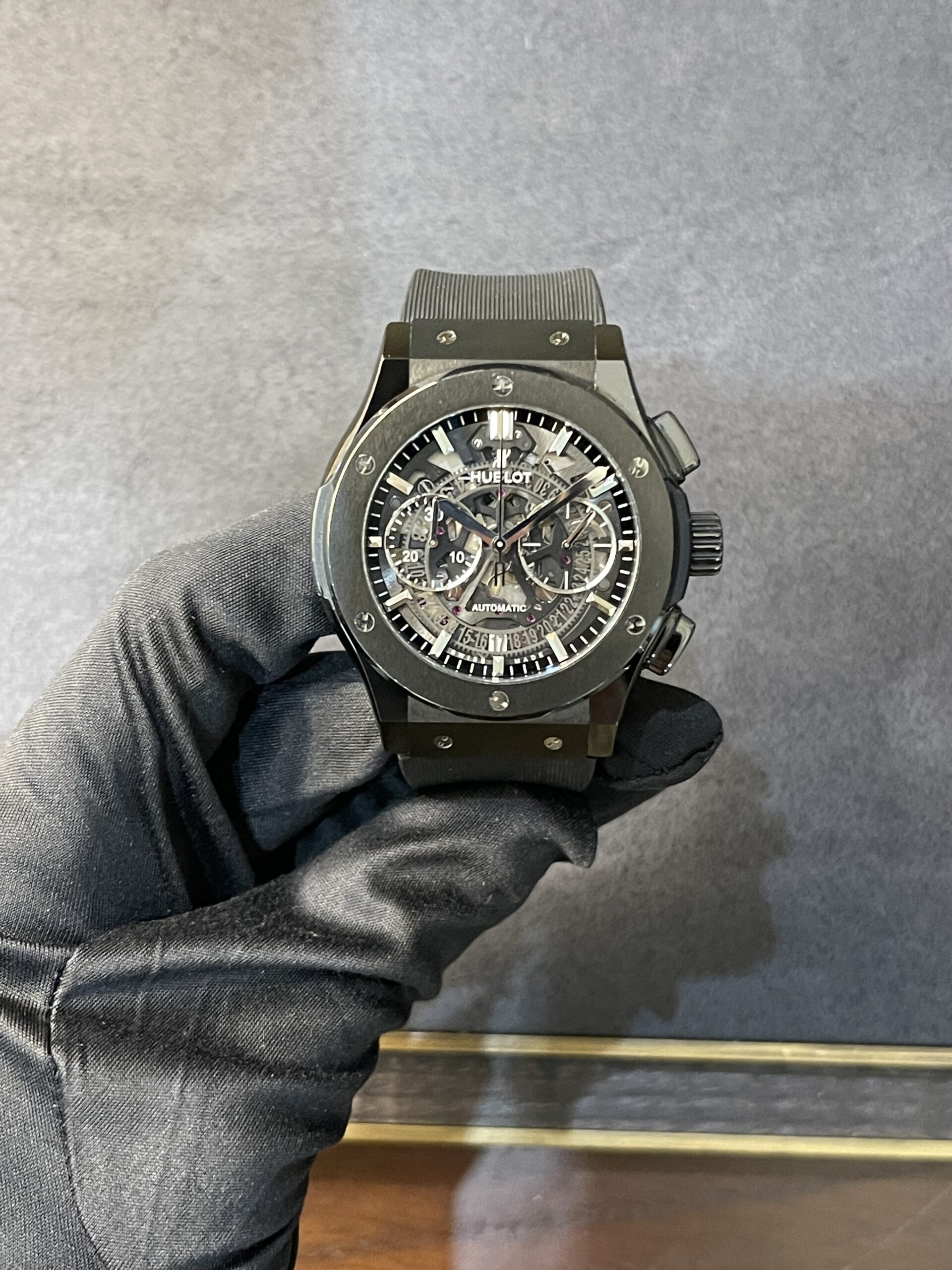 Skeleton-dial black Hublot automatic wristwatch on a gloved hand against a gray backdrop.