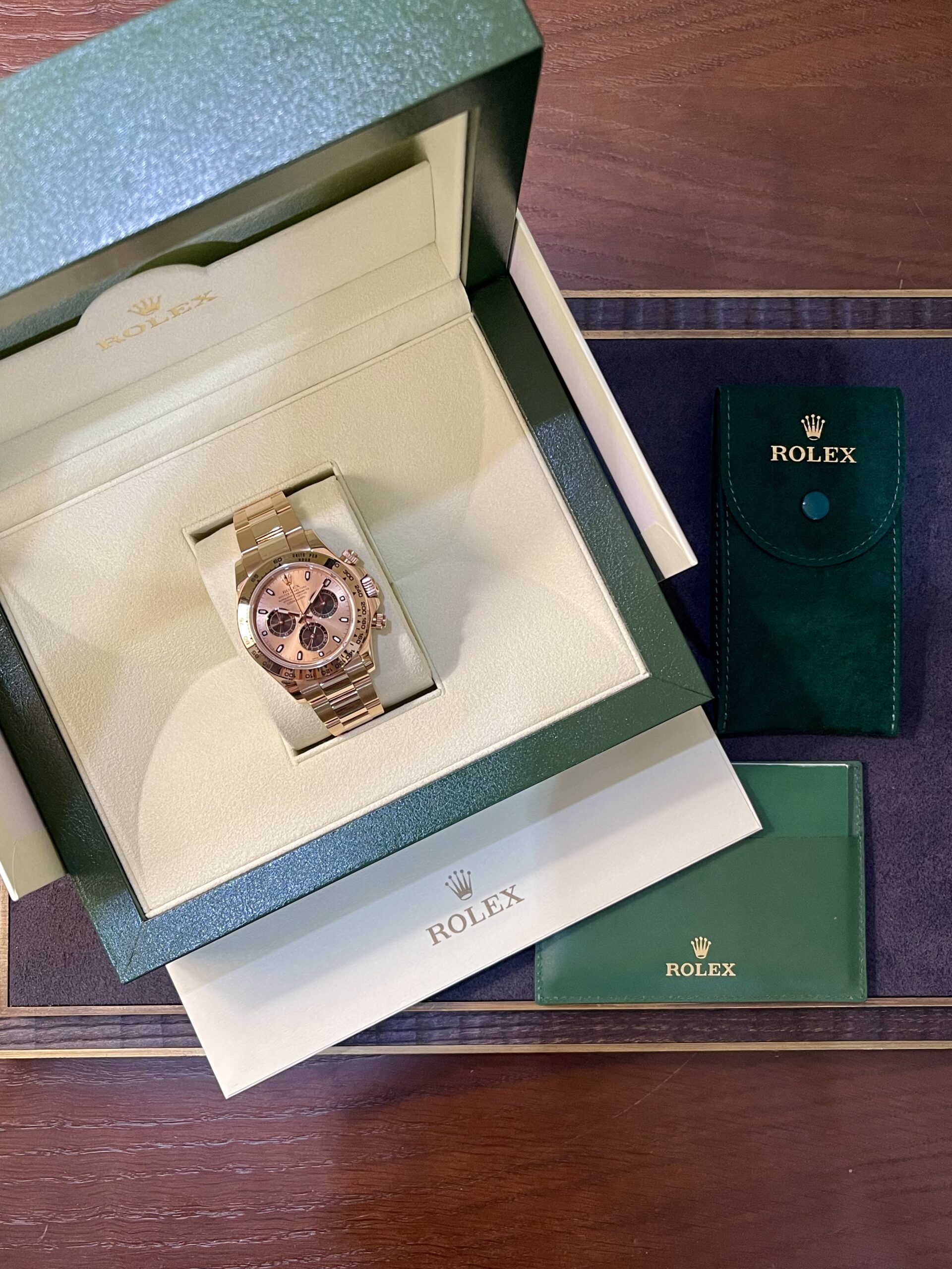 Rose gold Rolex watch in an open green presentation box with cream interior and matching green paperwork pouches nearby.
