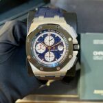 Close-up of an Audemars Piguet chronograph watch with a blue dial and white subdials on a display cushion. The blue strap and brushed metal case are visible.