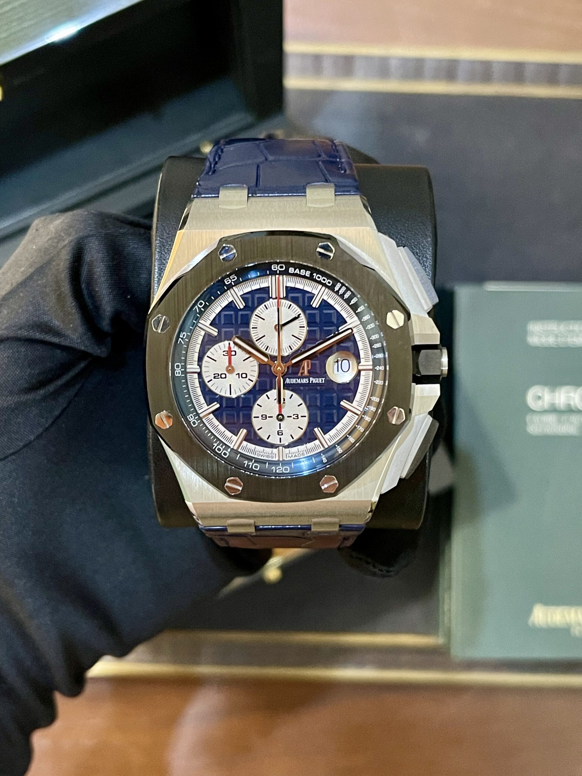 Close-up of an Audemars Piguet chronograph watch with a blue dial and white subdials on a display cushion. The blue strap and brushed metal case are visible.