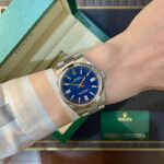 Wrist wearing a blue Rolex Oyster Perpetual with silver bracelet next to a green Rolex box and card box.