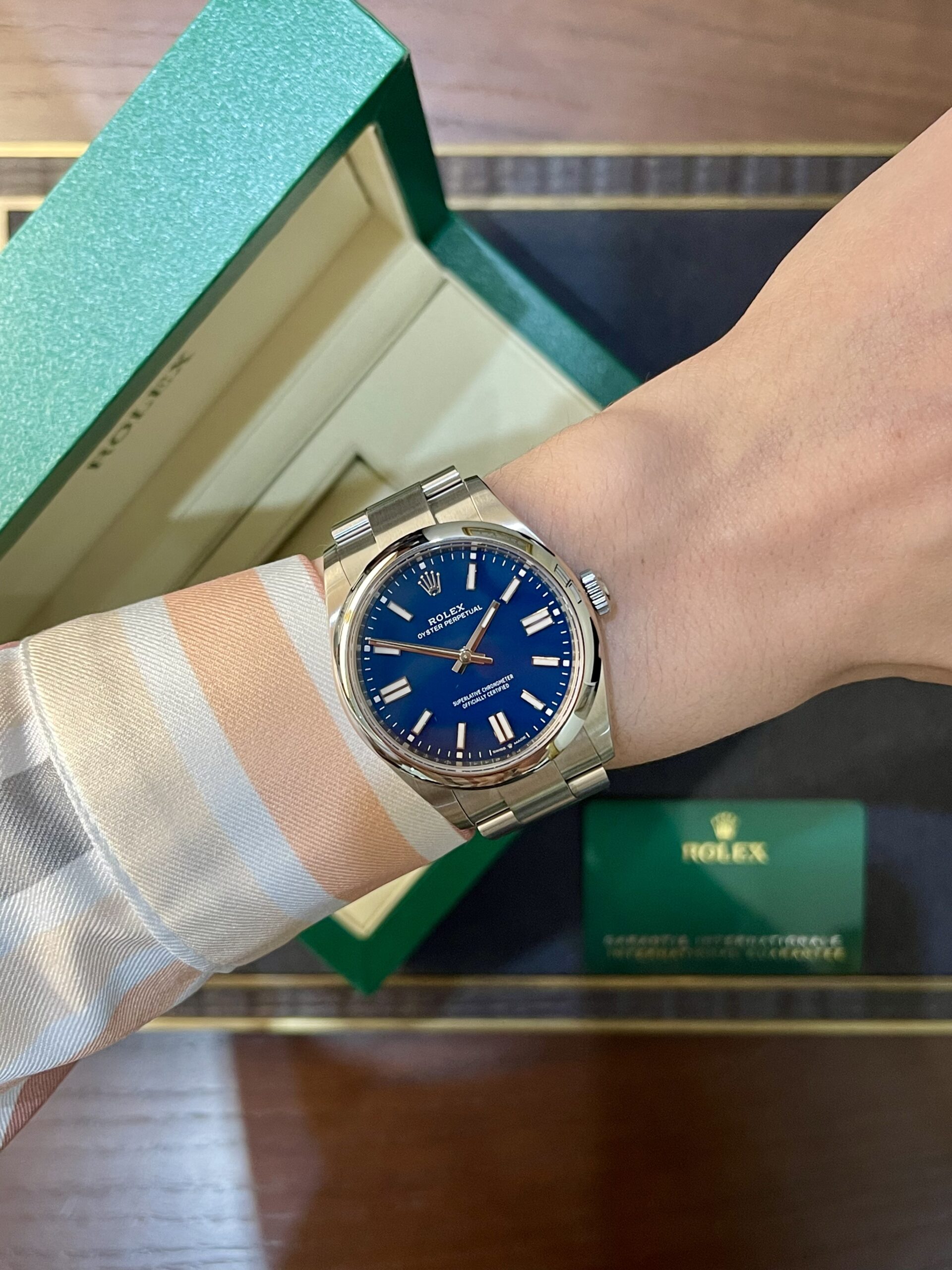 Wrist wearing a blue Rolex Oyster Perpetual with silver bracelet next to a green Rolex box and card box.