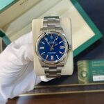 Rolex Oyster Perpetual with a blue dial in an open green box with a metal bracelet (watch on a beige cushion).