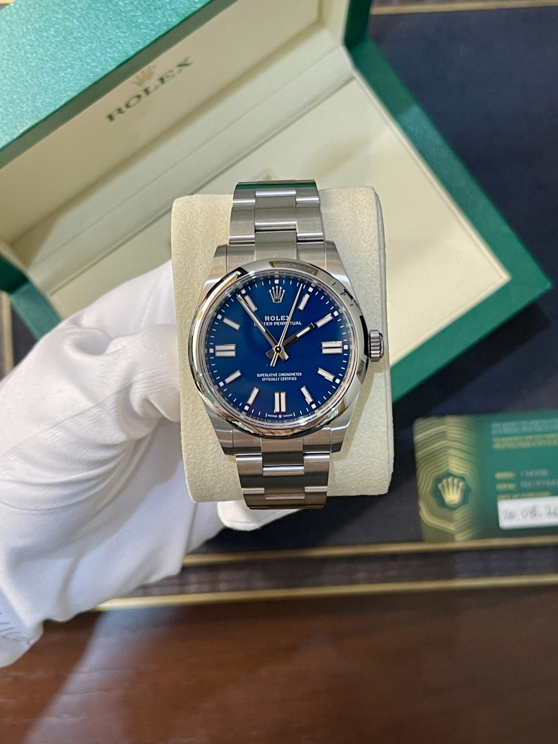 Rolex Oyster Perpetual with a blue dial in an open green box with a metal bracelet (watch on a beige cushion).
