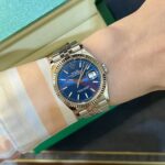 Wrist wearing a silver Rolex Datejust with a blue dial and magnified date window beside a green box, paired with a striped shirt cuff.