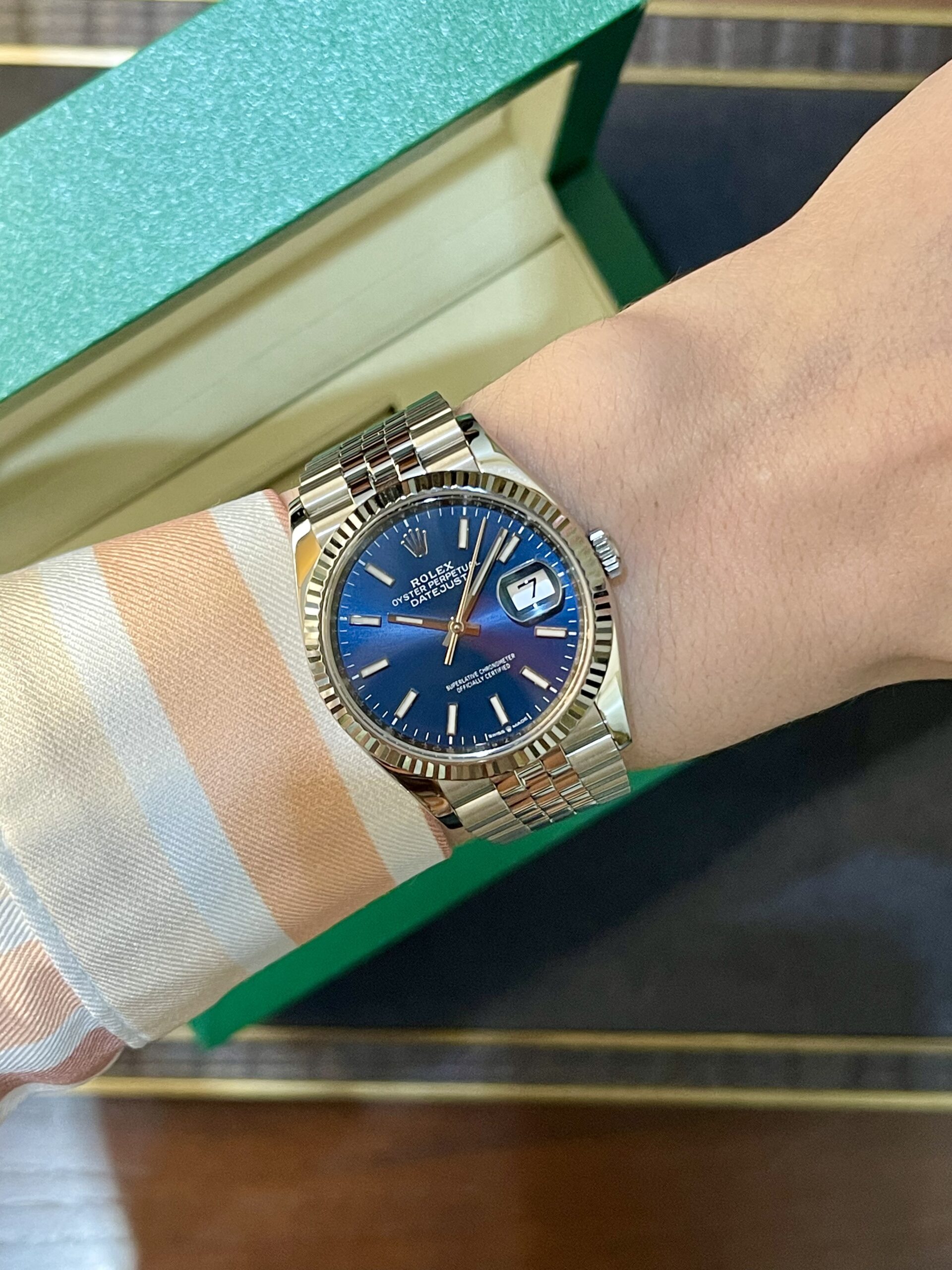 Wrist wearing a silver Rolex Datejust with a blue dial and magnified date window beside a green box, paired with a striped shirt cuff.