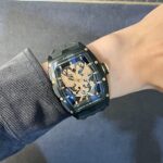 Close-up of a black Cvstos wristwatch with a gold dragon skeleton dial on a wrist with a ribbed sleeve and blue accents on the face.