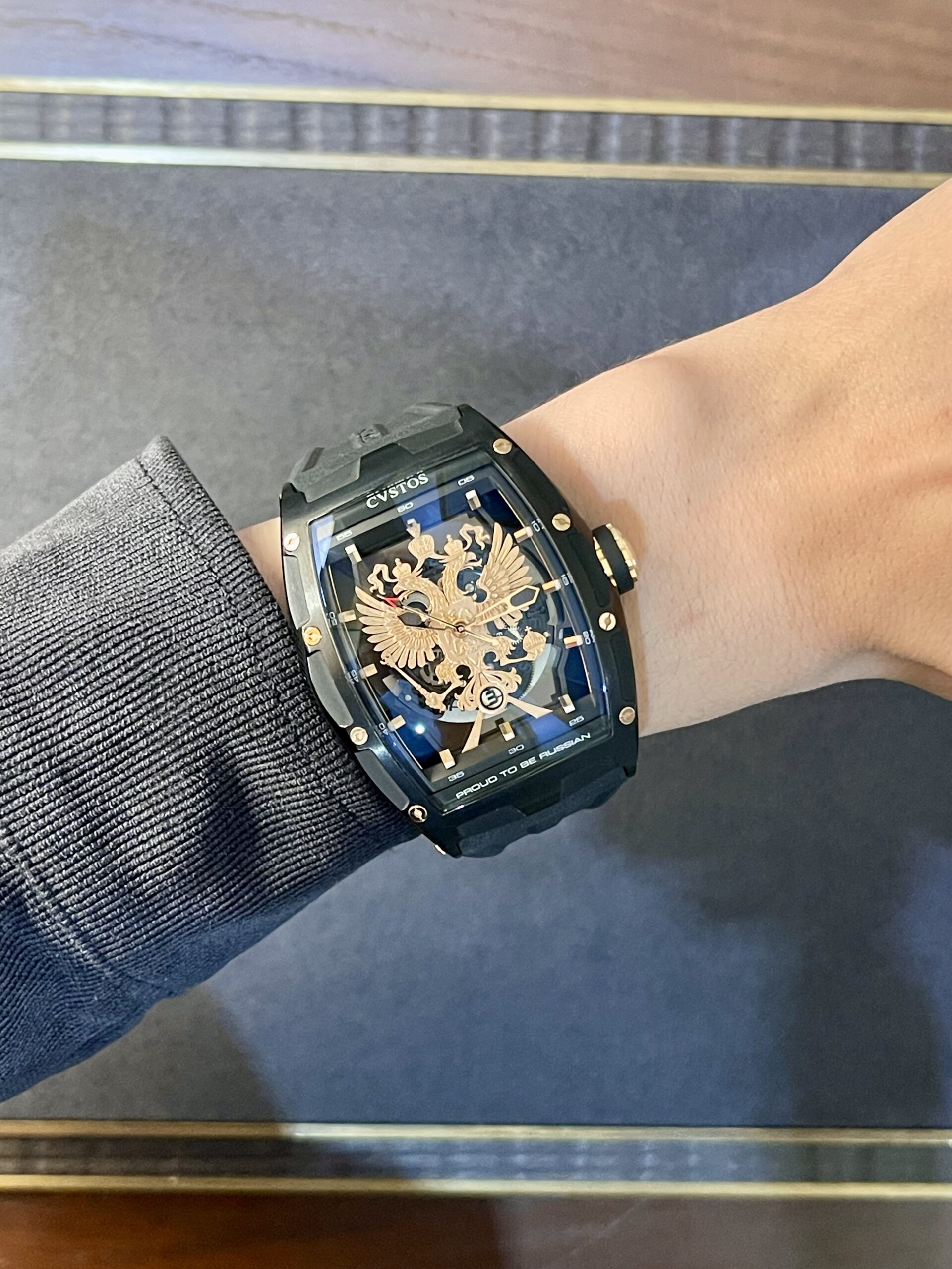 Close-up of a black Cvstos wristwatch with a gold dragon skeleton dial on a wrist with a ribbed sleeve and blue accents on the face.