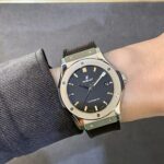 Wristwatch on a left forearm: stainless steel Hublot with black dial, gold hour markers, date window, and brown leather strap wearing over a gray sleeve.