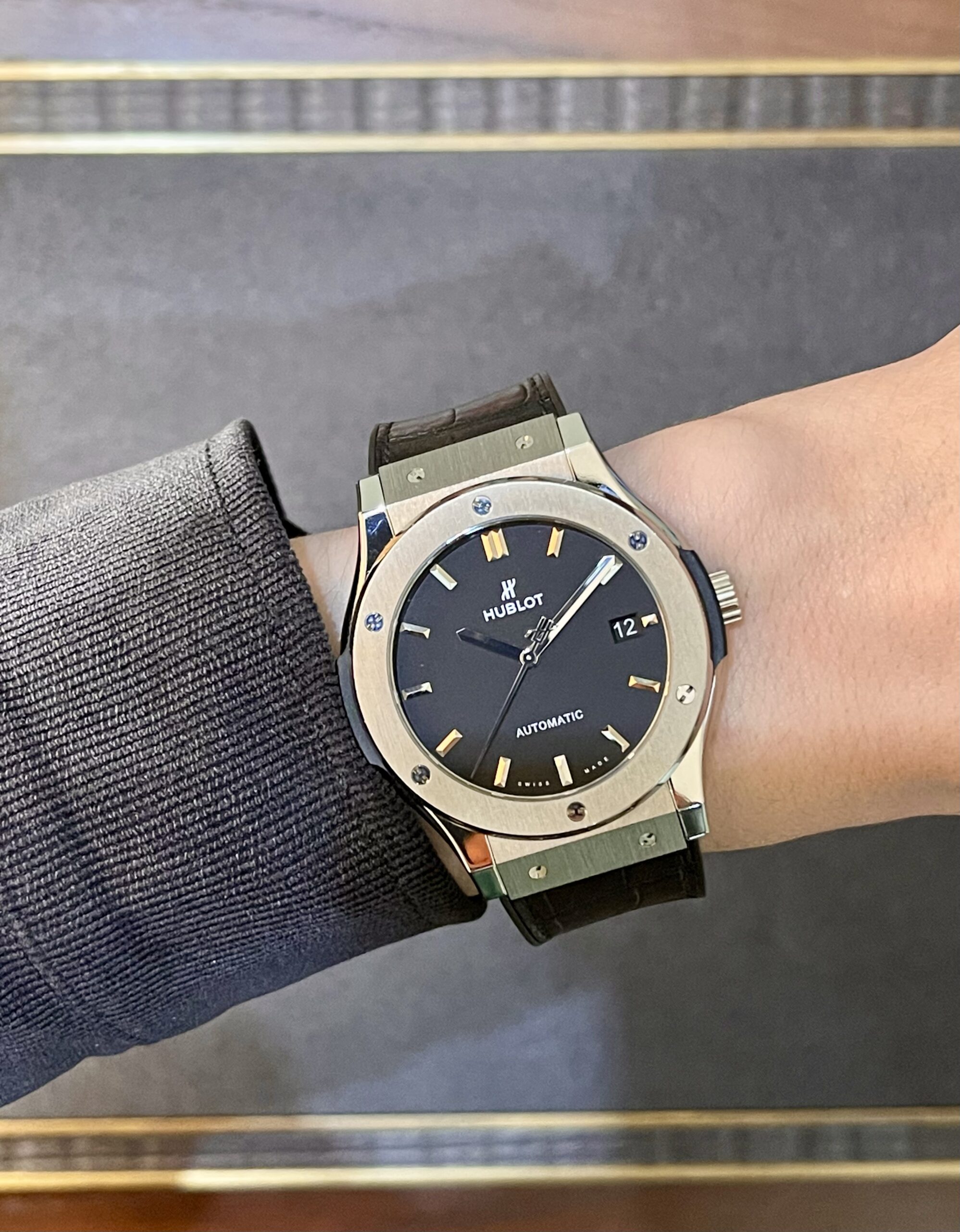 Wristwatch on a left forearm: stainless steel Hublot with black dial, gold hour markers, date window, and brown leather strap wearing over a gray sleeve.
