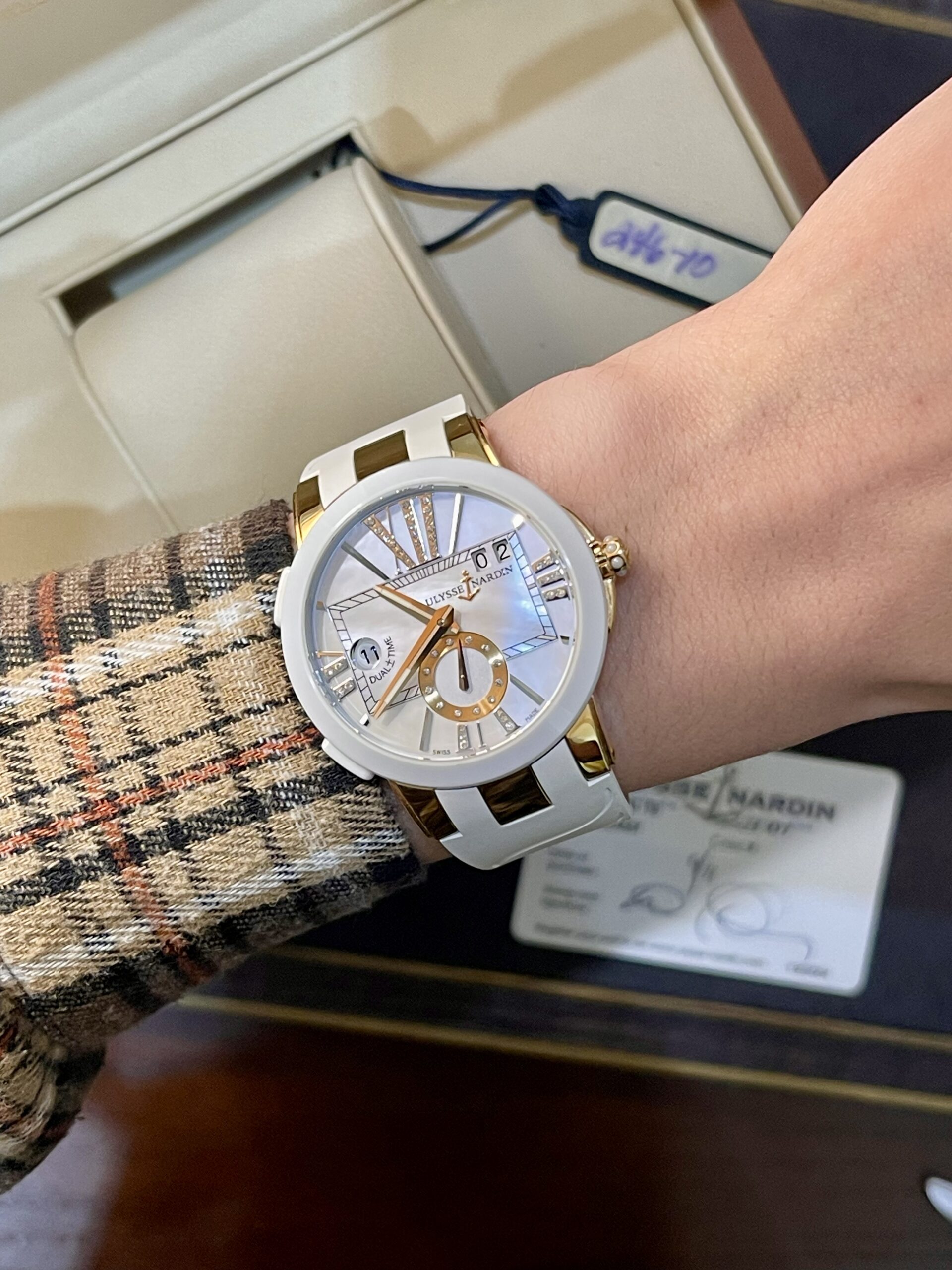 Wrist wearing a white and gold Ulysse Nardin watch with a mother-of-pearl dial and roman numerals, over a plaid sleeve