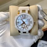 White and gold Ulysse Nardin wristwatch on a beige display cushion, showing a jeweled dial and 'Dual Time' indication (gloved hands holding it).