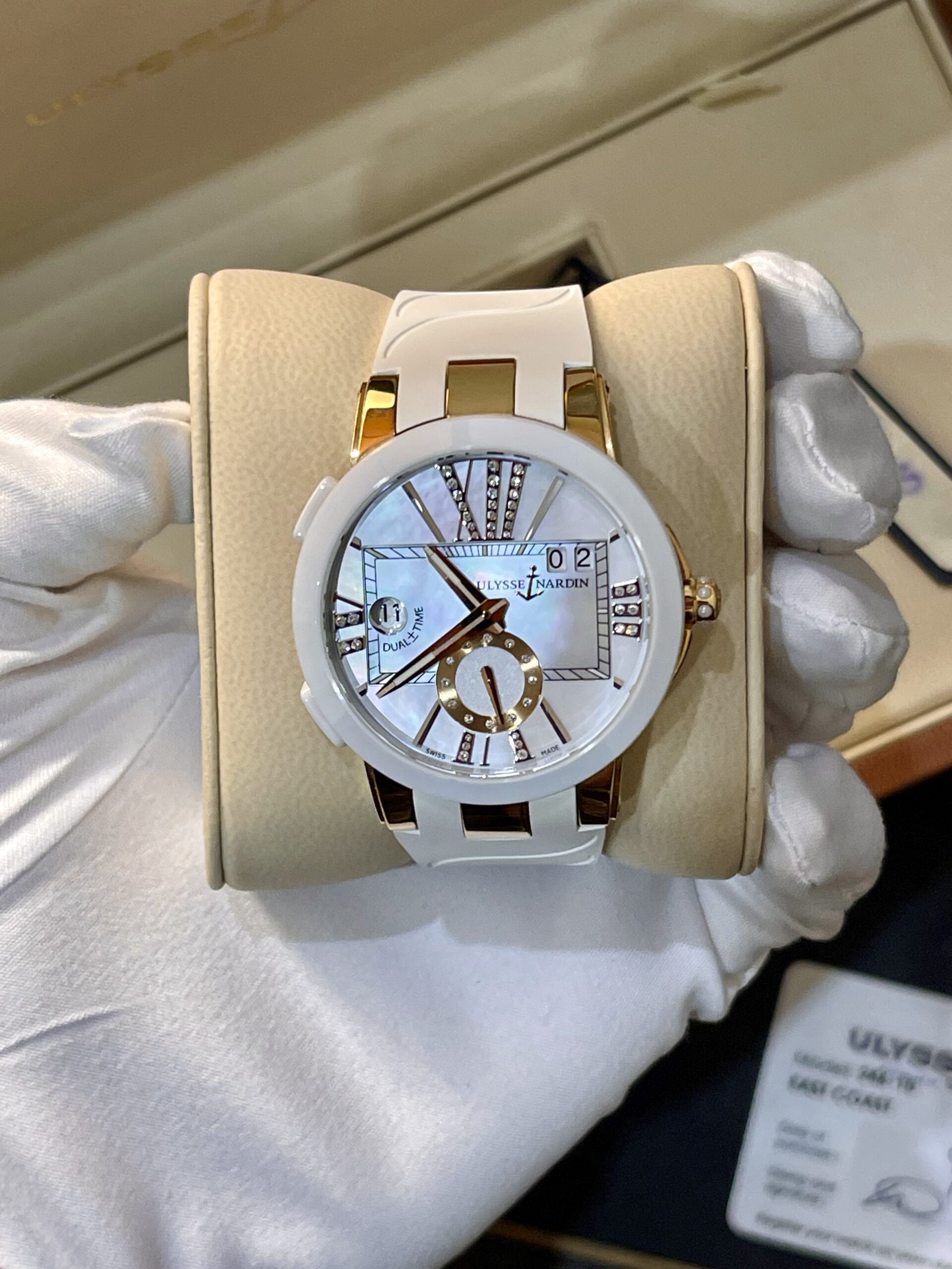 White and gold Ulysse Nardin wristwatch on a beige display cushion, showing a jeweled dial and 'Dual Time' indication (gloved hands holding it).