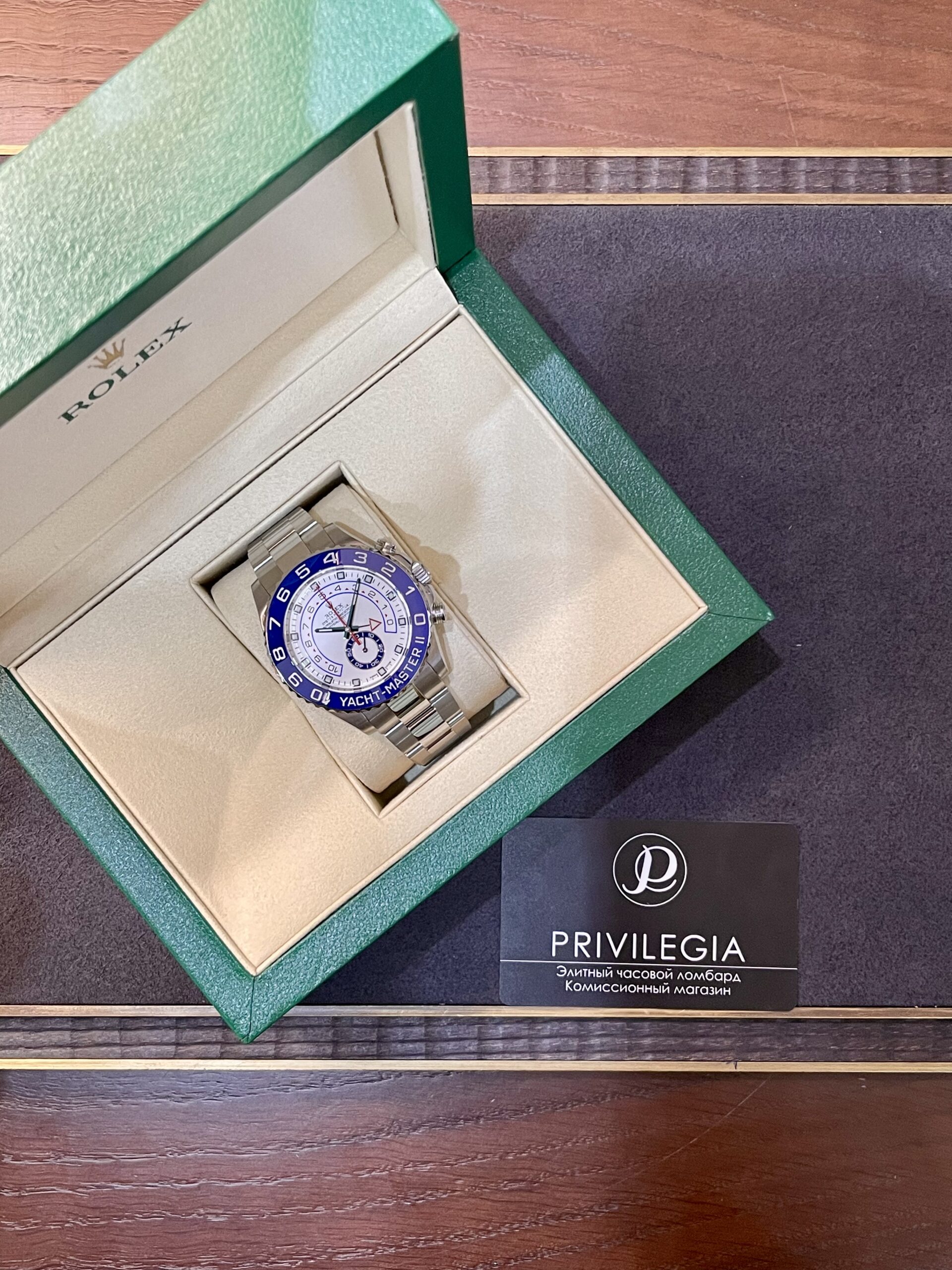 Opened green Rolex box displaying a silver Yacht-Master watch with a blue bezel on a beige insert; Privilegia card nearby.
