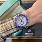 Rolex Yacht-Master II style watch on a plaid-sleeve arm; blue bezel, white dial, silver bracelet, with a green box in the background.