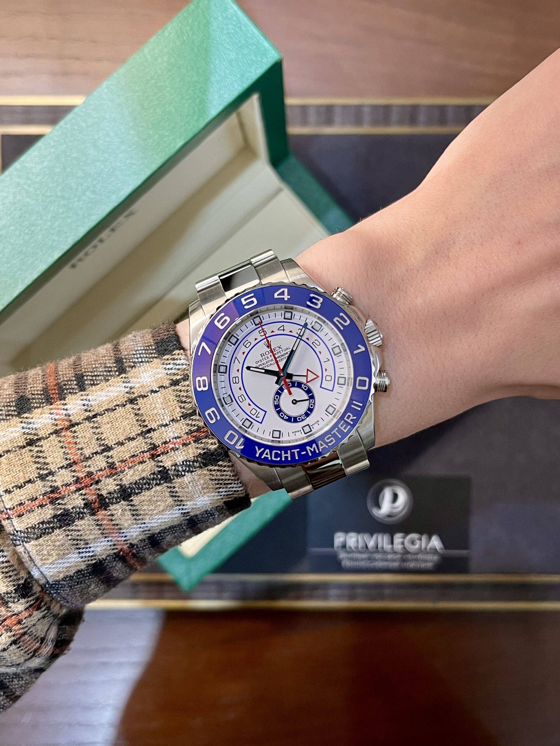 Rolex Yacht-Master II style watch on a plaid-sleeve arm; blue bezel, white dial, silver bracelet, with a green box in the background.