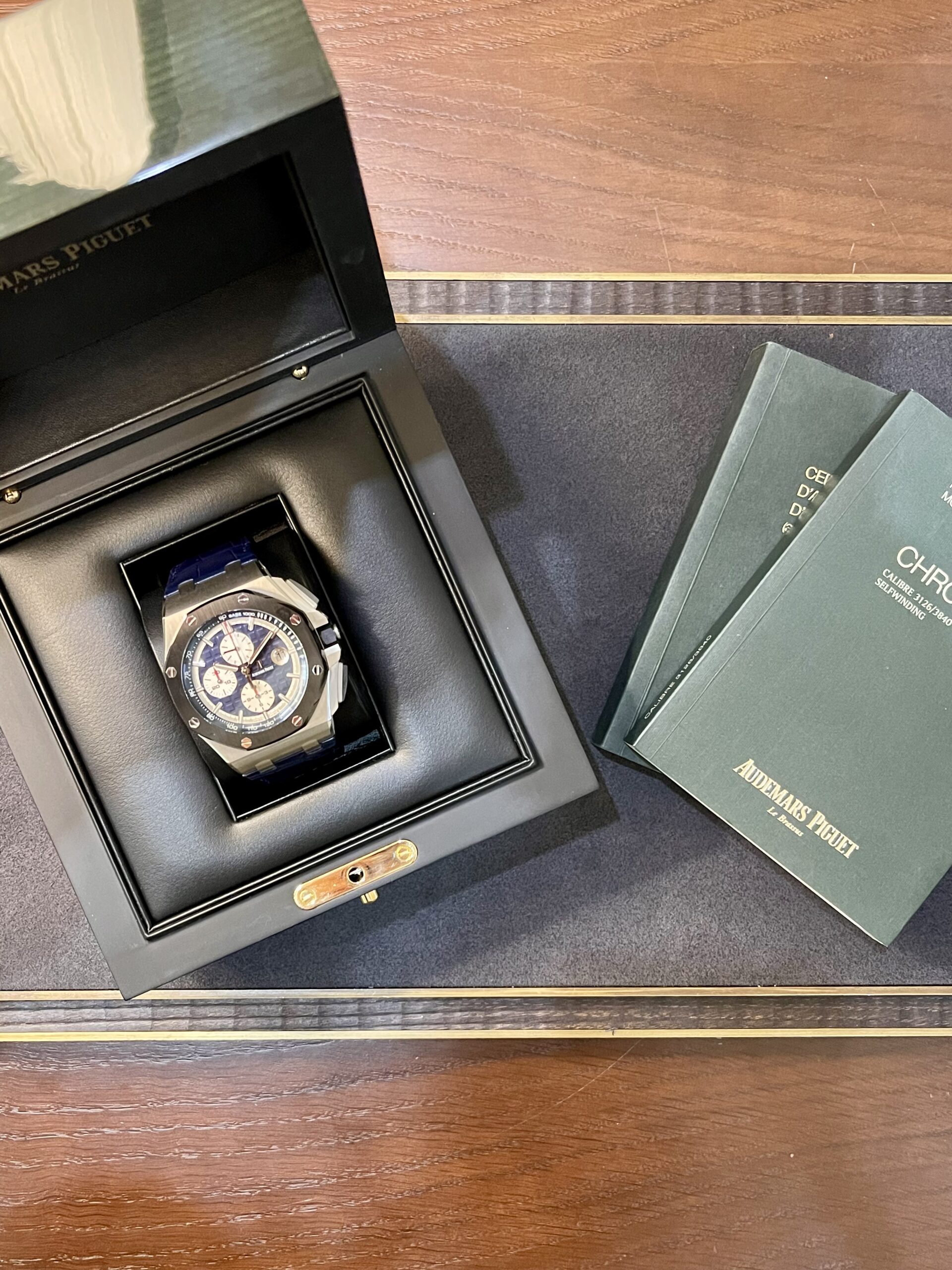 Luxurious Audemars Piguet watch in an open black presentation box with green manuals nearby on a wooden surface
