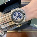 Wristwatch with a blue patterned dial on a metal bezel, worn over a plaid jacket sleeve, box in the background