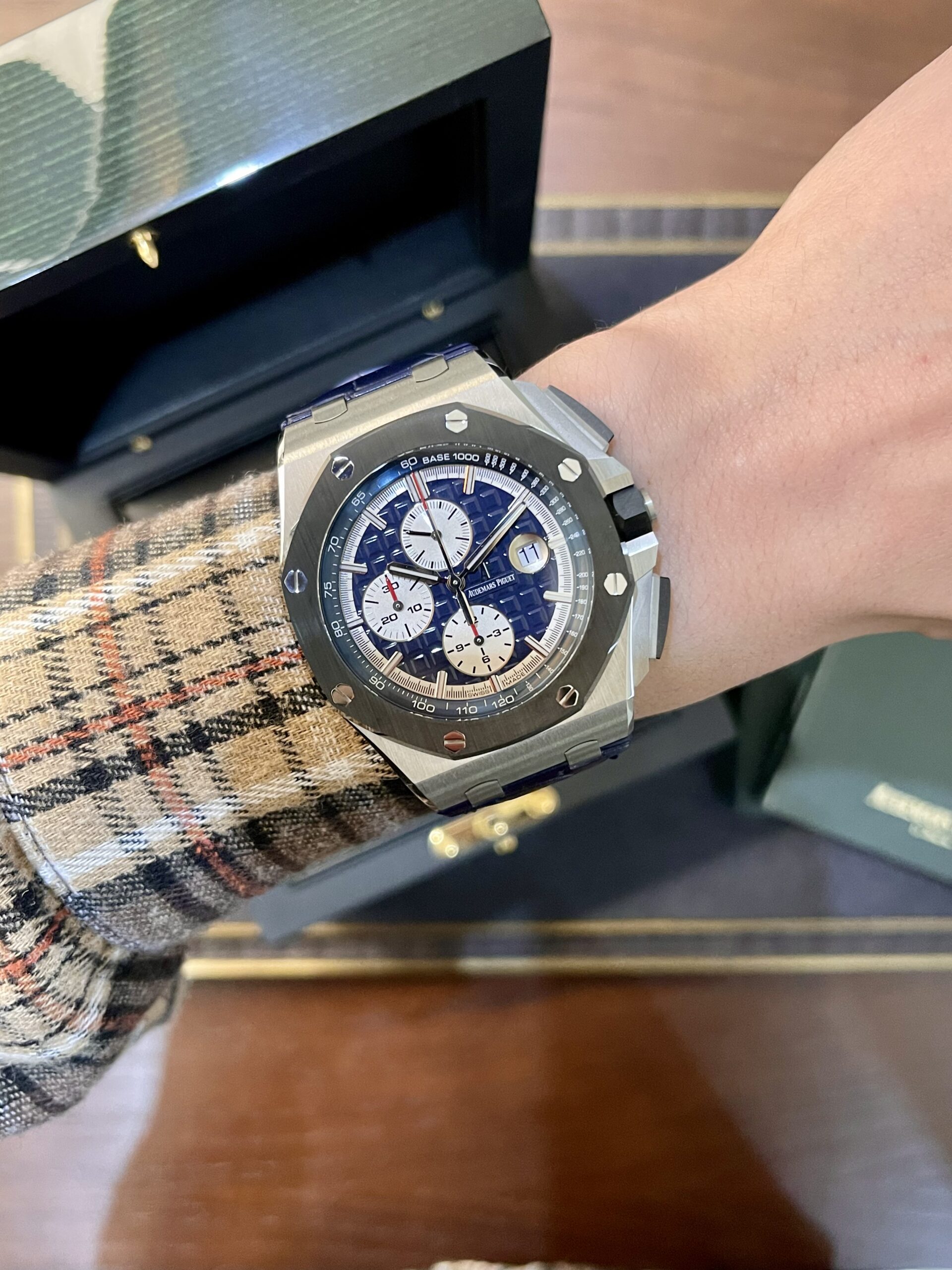 Wristwatch with a blue patterned dial on a metal bezel, worn over a plaid jacket sleeve, box in the background