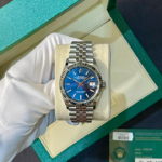 Rolex watch with blue dial and date display in a green presentation box held by gloved hand, showing stainless steel bracelet and fluted bezel.