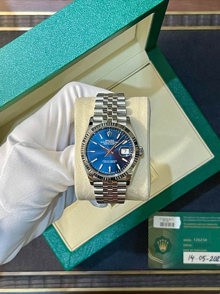Rolex watch with blue dial and date display in a green presentation box held by gloved hand, showing stainless steel bracelet and fluted bezel.