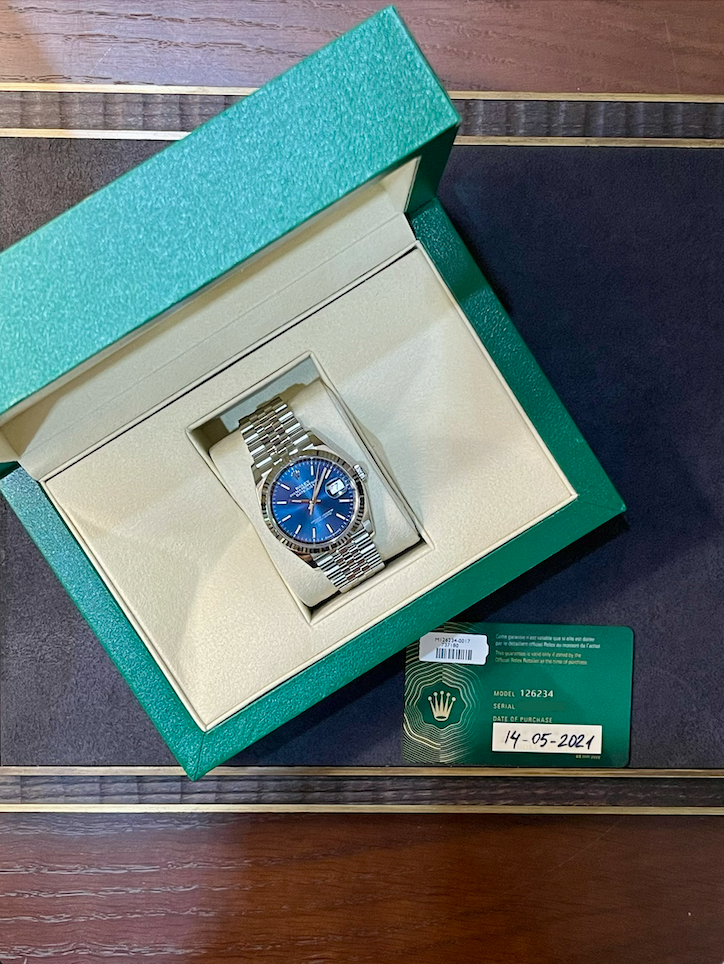 Rolex watch with a blue dial in a green presentation box and accompanying certificate showing 14-05-2021 date of purchase, stainless steel bracelet visible.