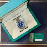 Rolex stainless steel watch with a blue dial in an open green presentation box, with a green warranty card nearby.